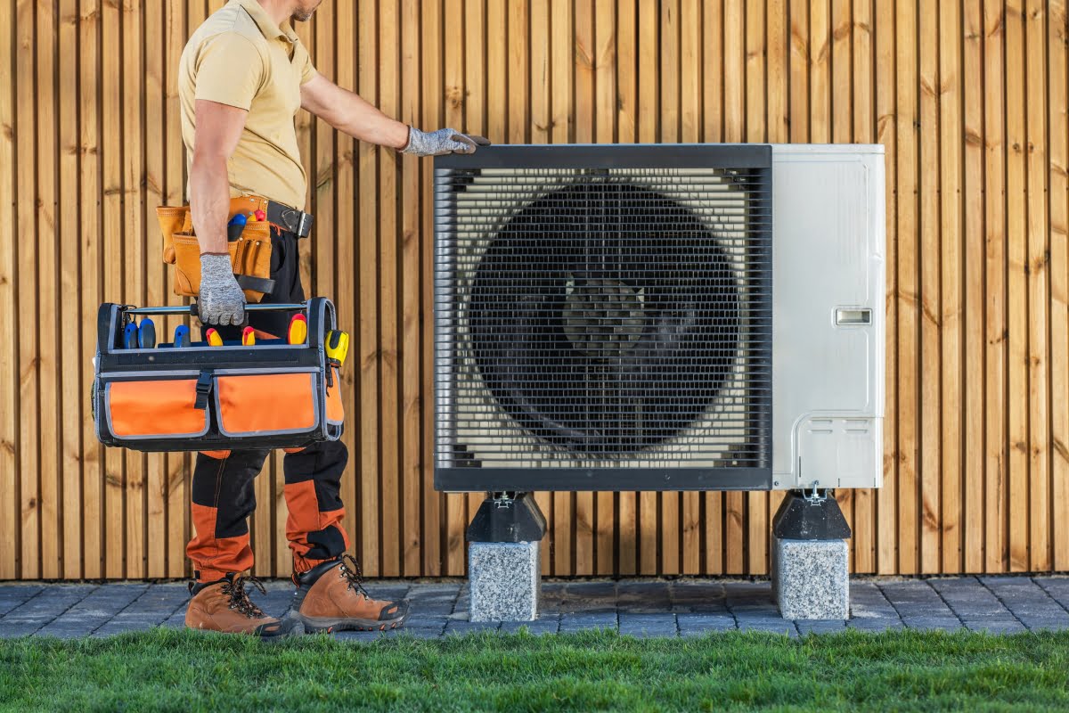 A technician in work attire holds a tool bag, checking an outdoor air conditioning unit mounted on a wooden wall, staying updated with the latest HVAC trends.