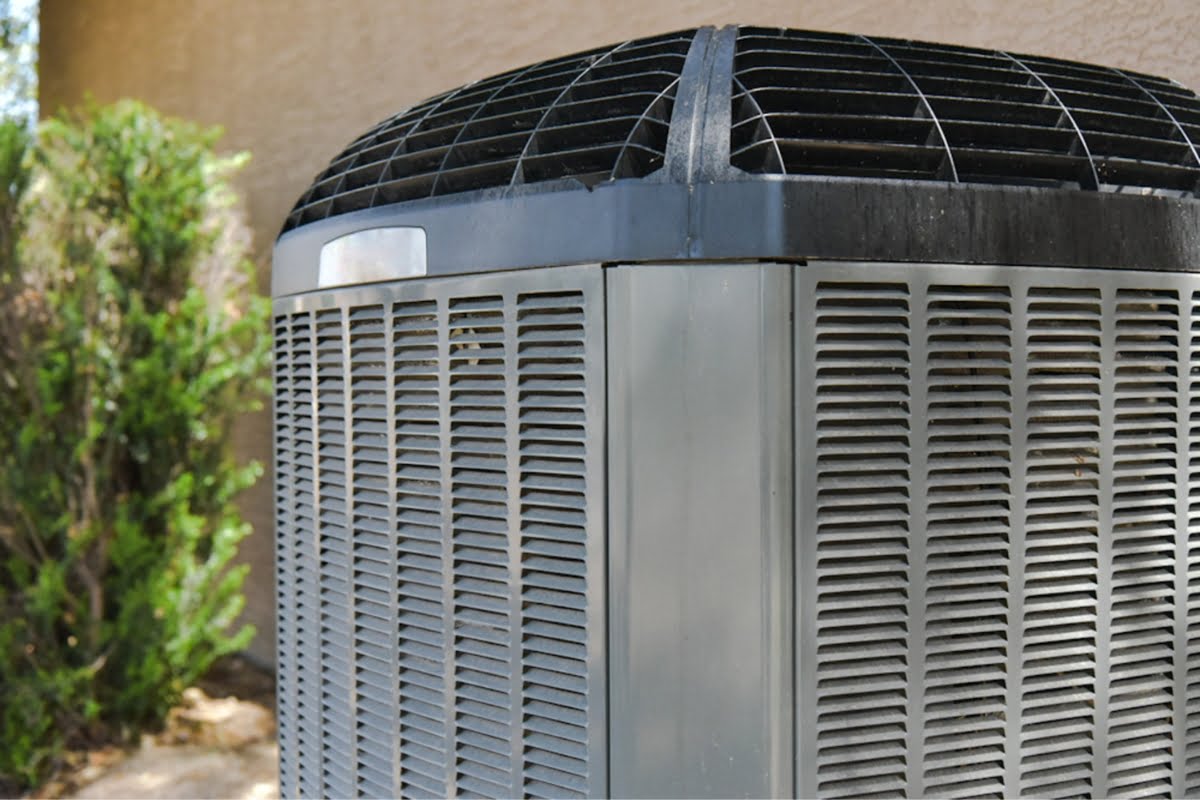 The outdoor air conditioning unit, featuring a metal grille and a sleek black top, is positioned next to a wall surrounded by greenery. This setup reflects the latest HVAC trends in both functionality and design.