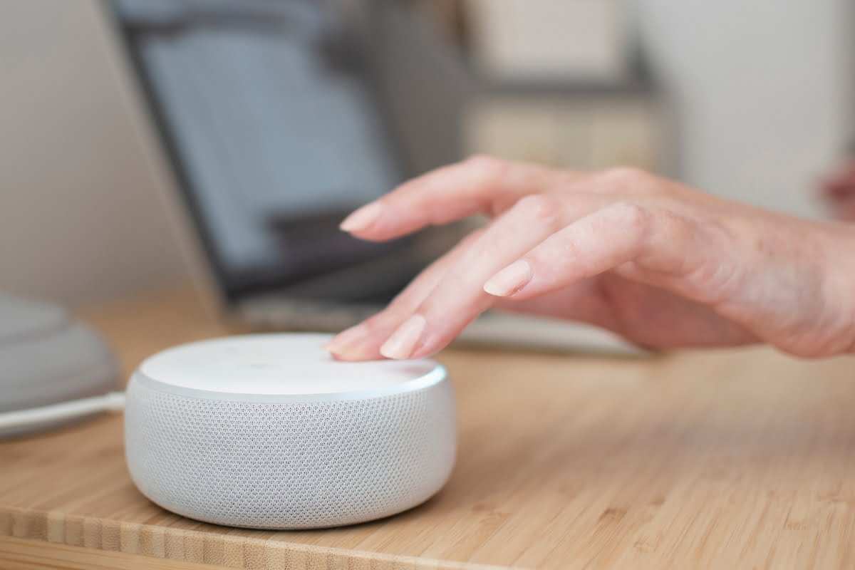 A person’s hand is about to touch a smart speaker placed on a desk next to an open laptop, possibly ready to inquire about the latest HVAC trends.