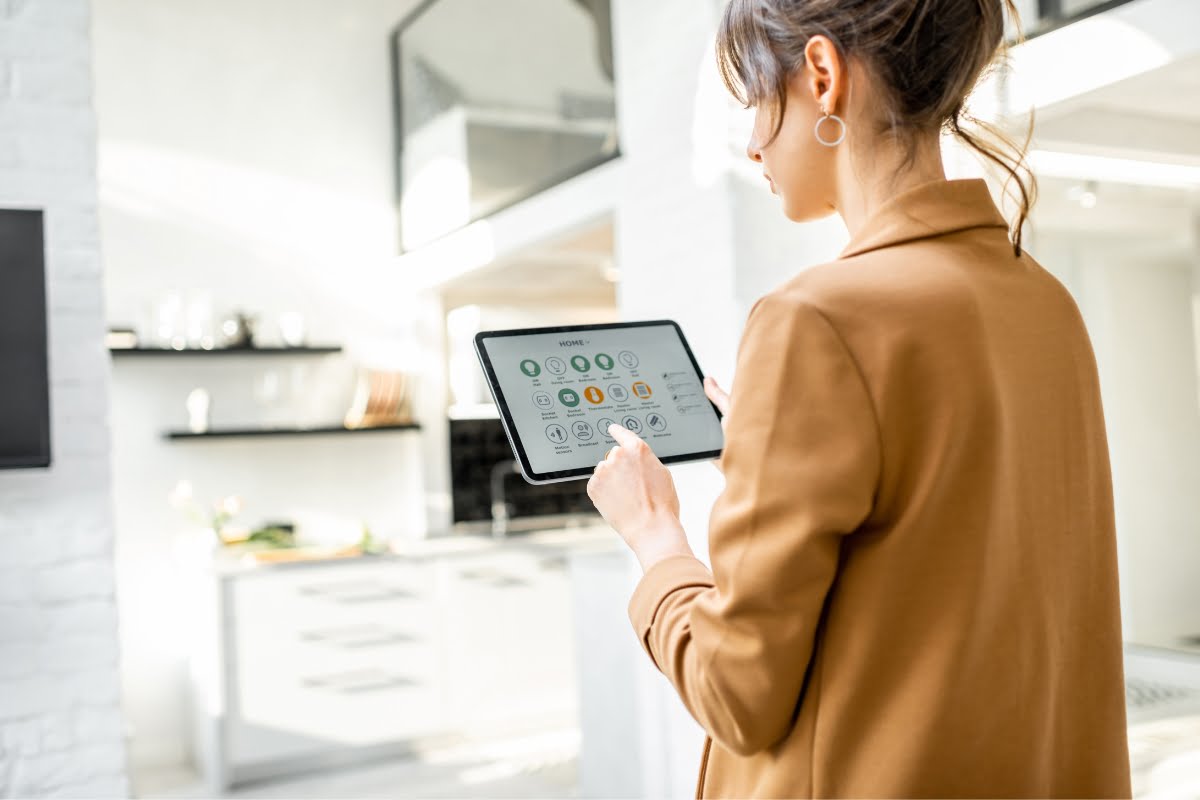 A person in a brown jacket uses a tablet in a modern kitchen. The tablet screen displays various icons, possibly for smart home and HVAC control trends. The kitchen is bright with white decor and open shelving.