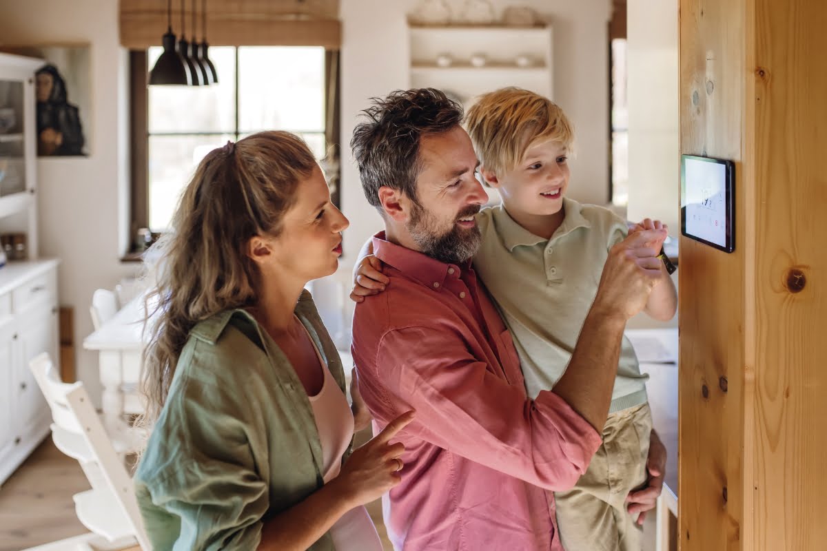 A man with a beard and a young boy adjust settings on a wall-mounted touchscreen, keeping up with the latest HVAC trends, while a woman with long hair stands next to them in a brightly lit kitchen.