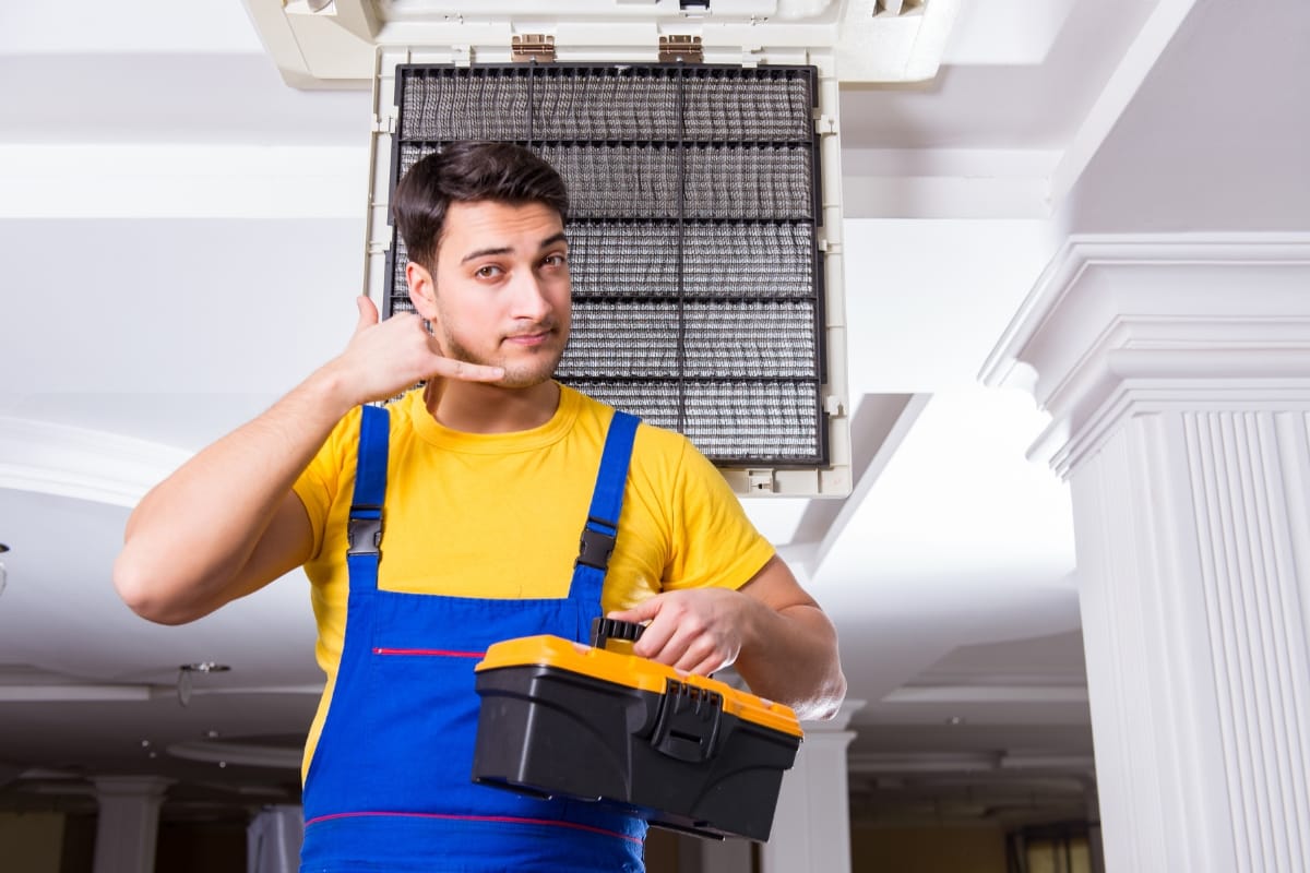 A technician in a yellow shirt and blue overalls holds a toolbox, gesturing with his hand beneath an open ceiling air conditioning unit, demonstrating HVAC fall maintenance techniques.