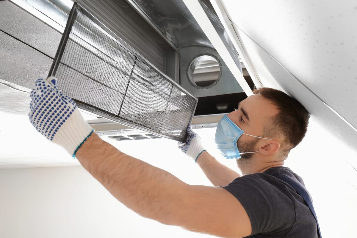 Wearing a mask and gloves, the technician expertly installs an air filter in the ceiling ventilation system as part of essential HVAC fall maintenance.