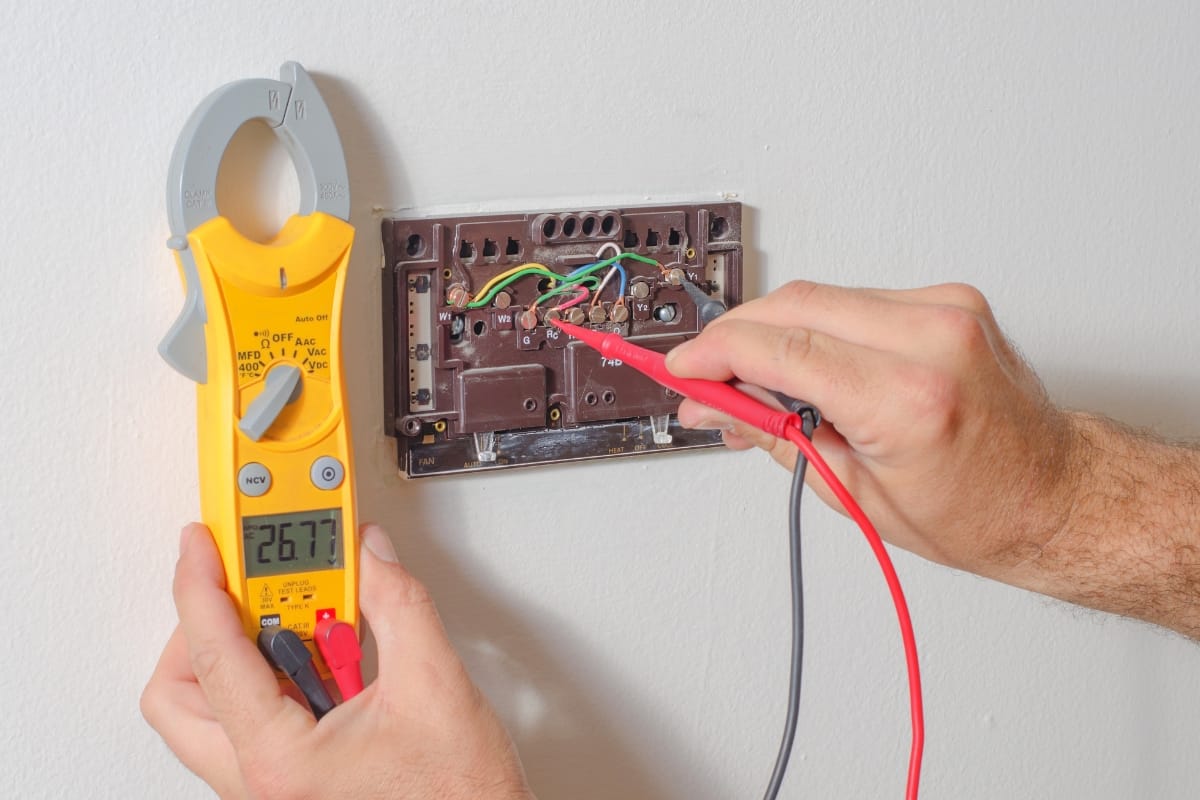 Hands are using a multimeter to test voltage on exposed electrical wiring within a wall socket as part of HVAC fall maintenance, ensuring systems run efficiently before the cold sets in.