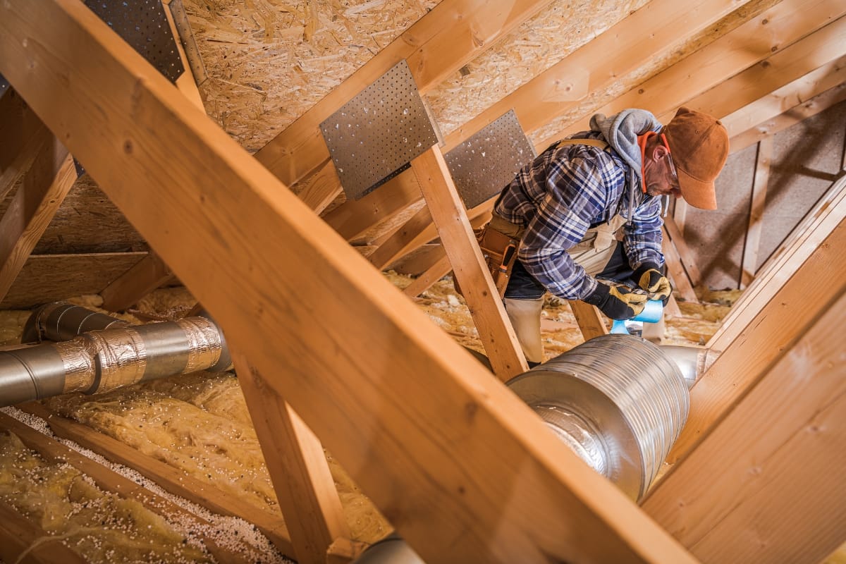 A person in work attire conducts HVAC repairs in an attic, surrounded by wooden beams and insulation as part of essential fall maintenance.