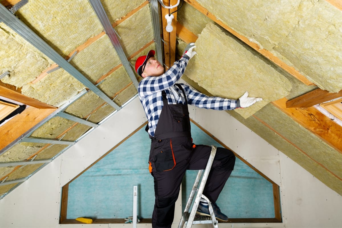 Person in checkered shirt and overalls installing insulation in an attic, standing on a ladder and adjusting a large section of insulating material as part of essential HVAC fall maintenance.