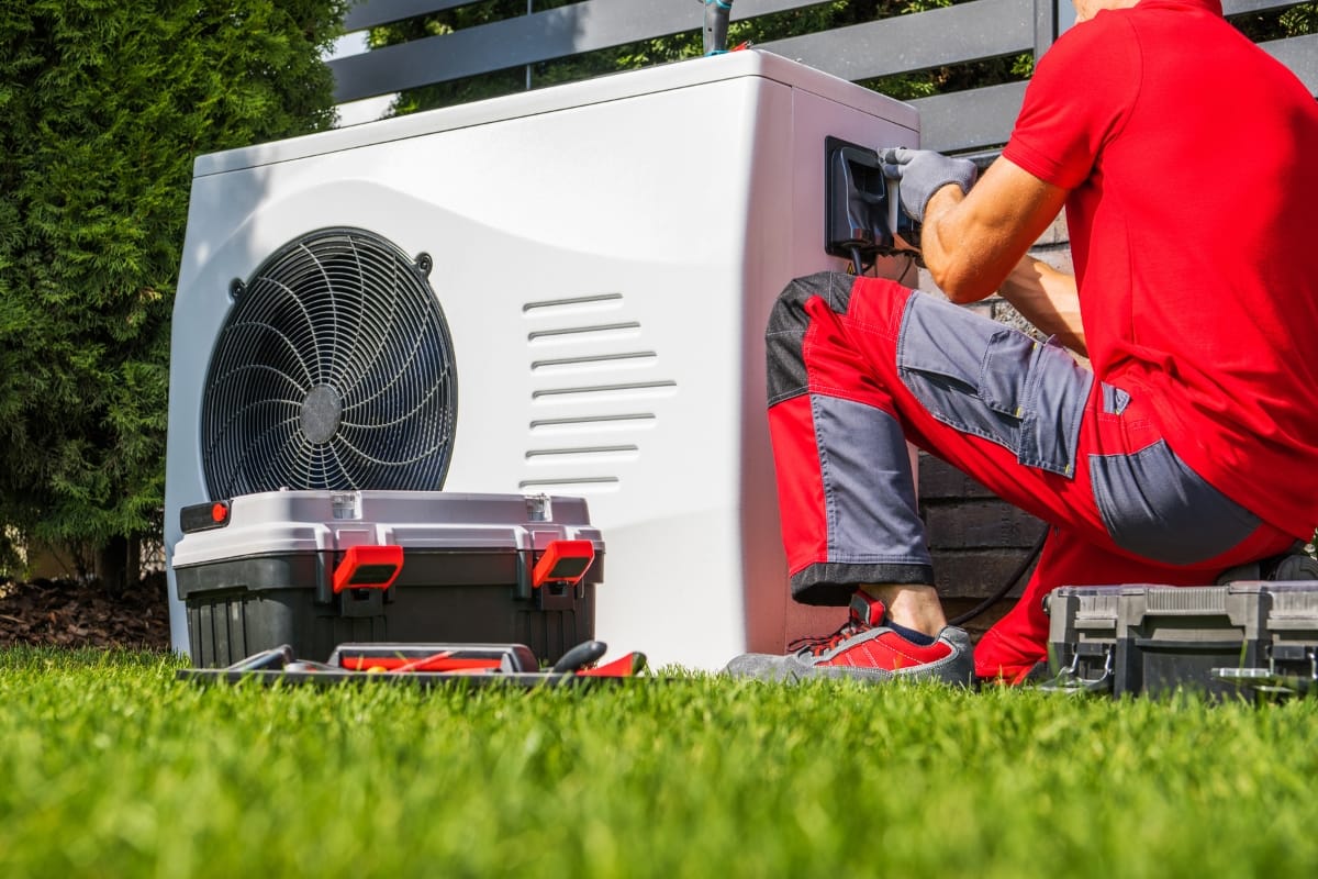 A technician in a red uniform diligently performs HVAC fall maintenance on an outdoor unit, surrounded by tools and equipment scattered across the grass.