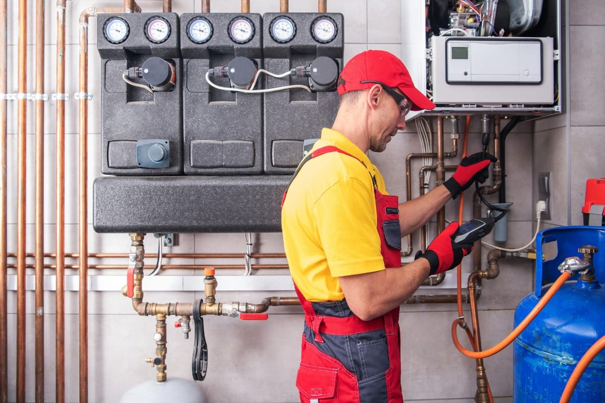 A technician in red overalls and cap checks gauges and pipes in a utility room, conducting HVAC fall maintenance with precision, holding a measurement device.