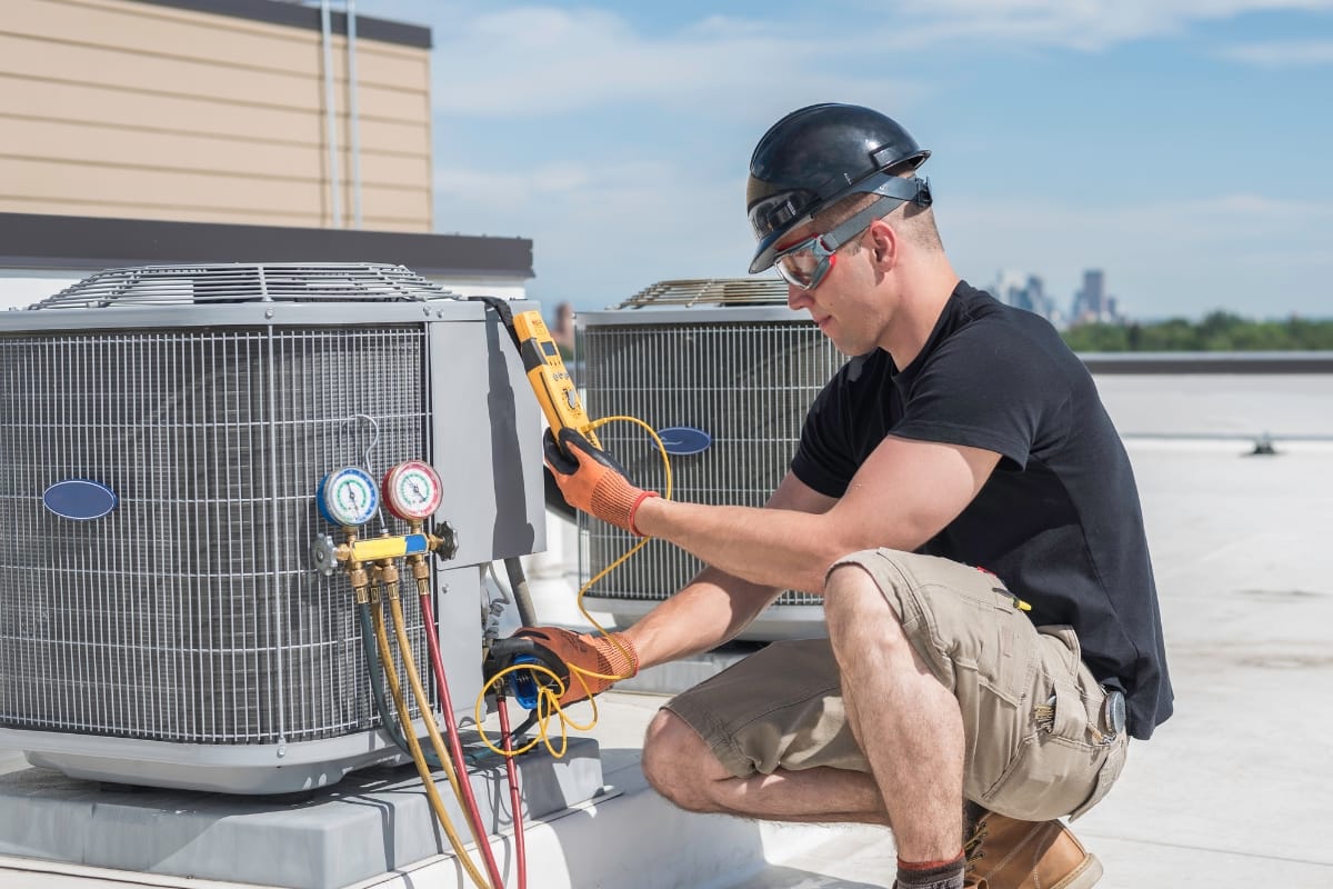 Technician in safety gear checks rooftop smart HVAC system with a digital multimeter on a sunny day.