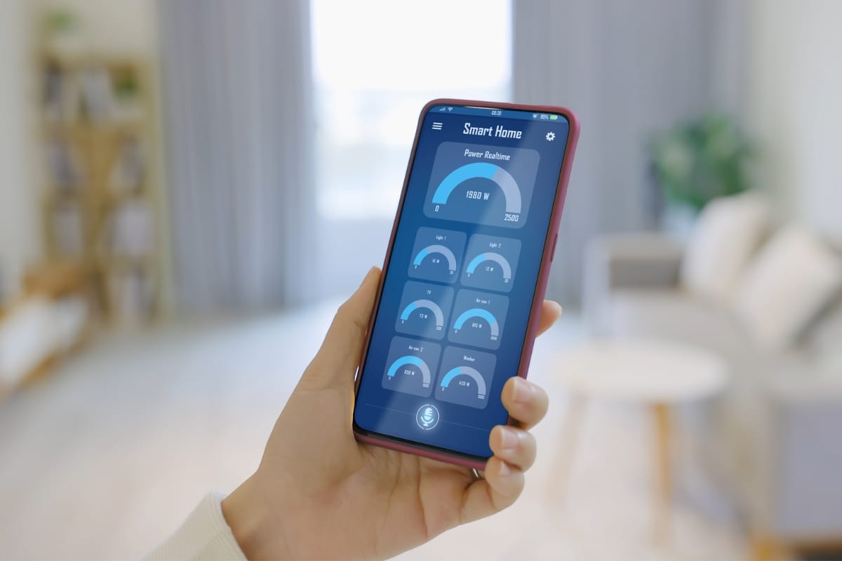 A hand holds a smartphone displaying a smart home app interface with various power usage metrics, including smart HVAC systems data, in a cozy living room setting.