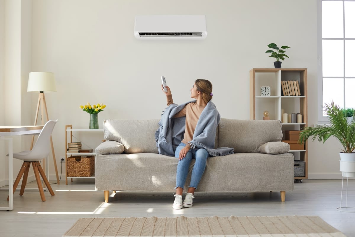 A woman lounging on a sofa uses a remote control to adjust a wall-mounted air conditioner, seamlessly integrated into the bright, modern living room's smart HVAC system.