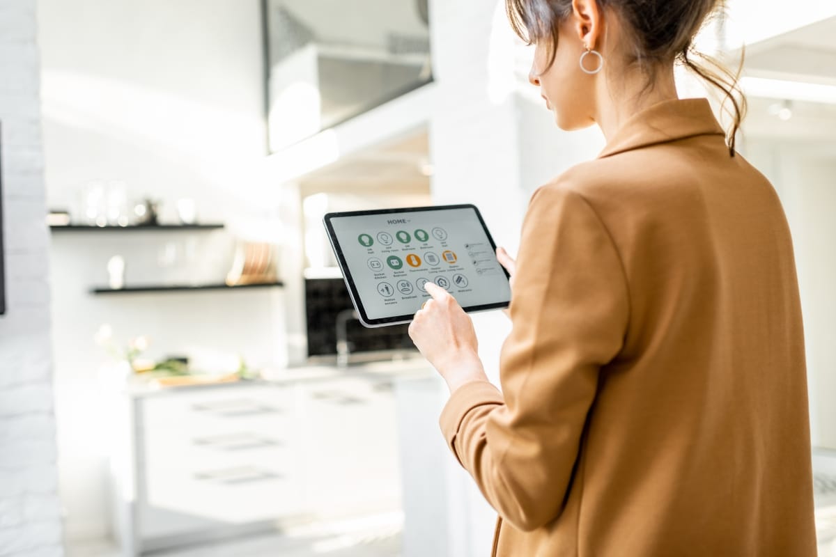 A woman in a brown jacket uses a tablet with a smart home interface, effortlessly managing the modern kitchen's smart HVAC systems.