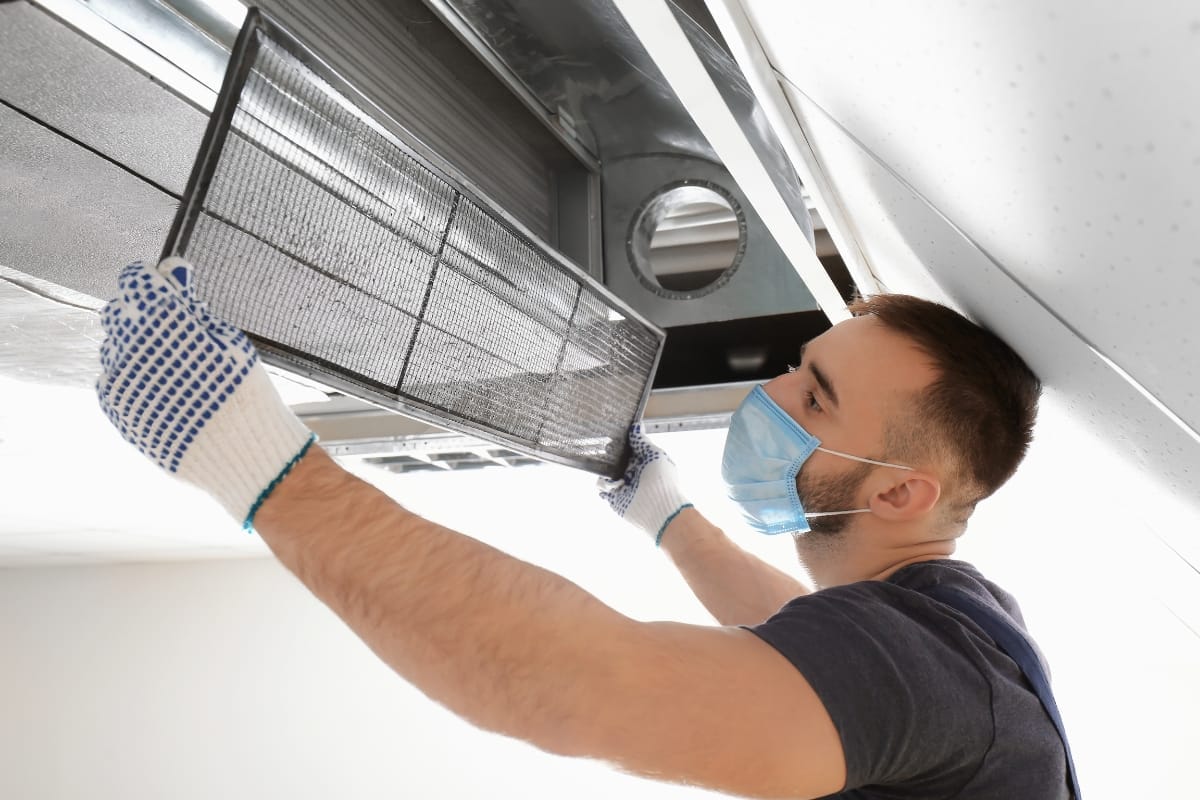 A person wearing gloves and a mask is installing or cleaning an air vent filter on a ceiling, following essential HVAC maintenance tips for improved air quality.