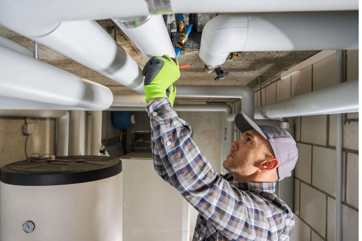 A man wearing gloves and a cap carefully inspects and adjusts a pipe in a utility room, showcasing essential HVAC maintenance tips.