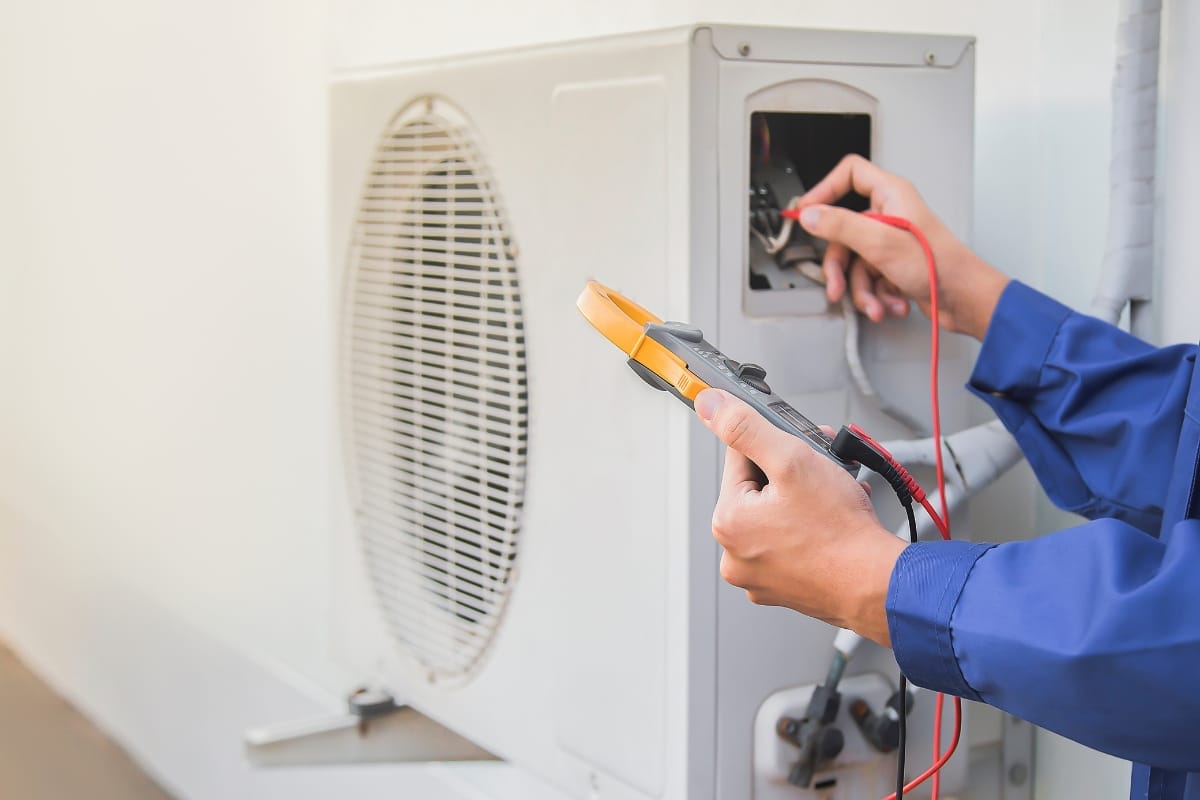 A technician in a blue uniform uses a multimeter to check an outdoor air conditioning unit, following essential HVAC maintenance tips to ensure efficient performance.