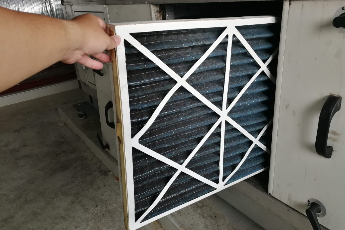 A hand is pulling a dirty air filter from an HVAC unit, highlighting dust accumulation as a reminder of essential HVAC maintenance tips. The filter features a white frame with a crisscross pattern, emphasizing the need for regular checking and cleaning.