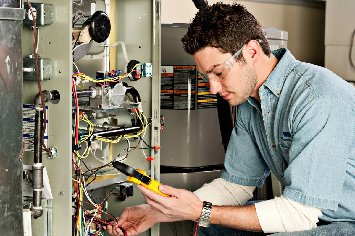 A person wearing protective eyewear uses a multimeter to inspect a complex array of wires in an HVAC system, searching for common furnace problems.