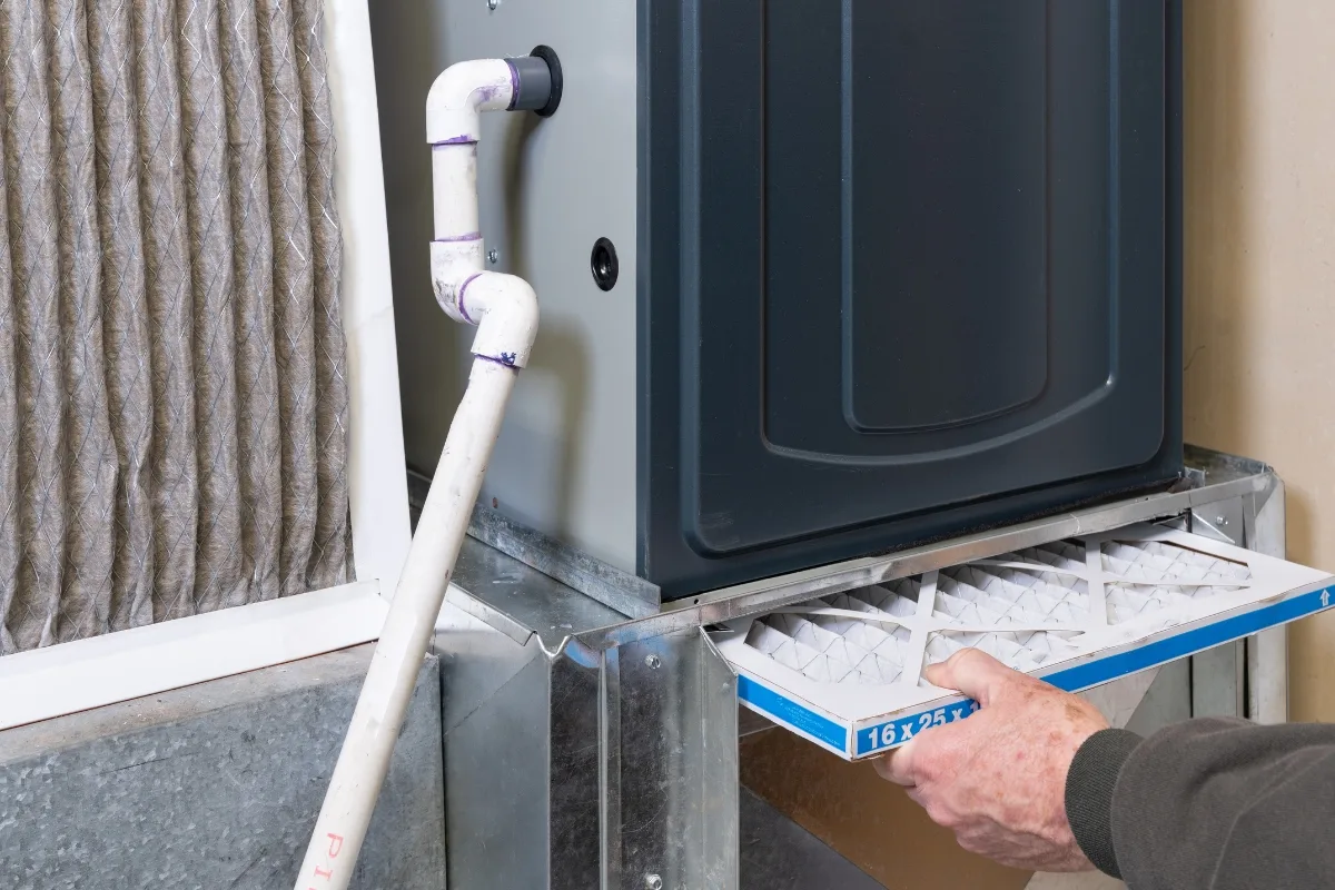 A person is replacing an air filter in a residential HVAC system, carefully positioned next to a wall and pipe, possibly addressing underlying heat pump issues for improved efficiency.
