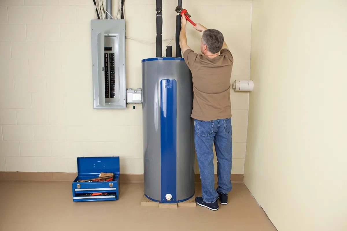 A man in a brown shirt uses a wrench to address heat pump issues, adjusting pipes on top of a blue and gray water heater next to an electrical panel. An open toolbox sits on the floor.