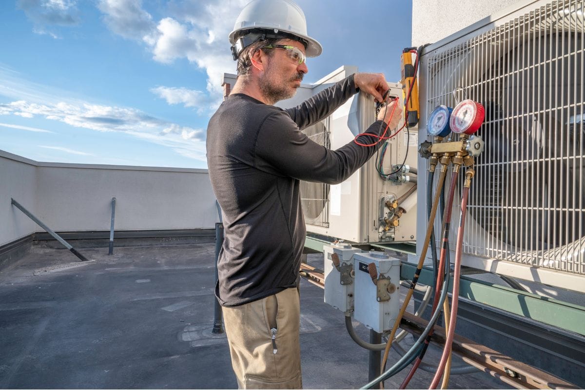 A technician in a hard hat and safety glasses checks HVAC equipment on a rooftop using a multimeter, with gauges and hoses clearly visible, showcasing expert HVAC services in Cumming.