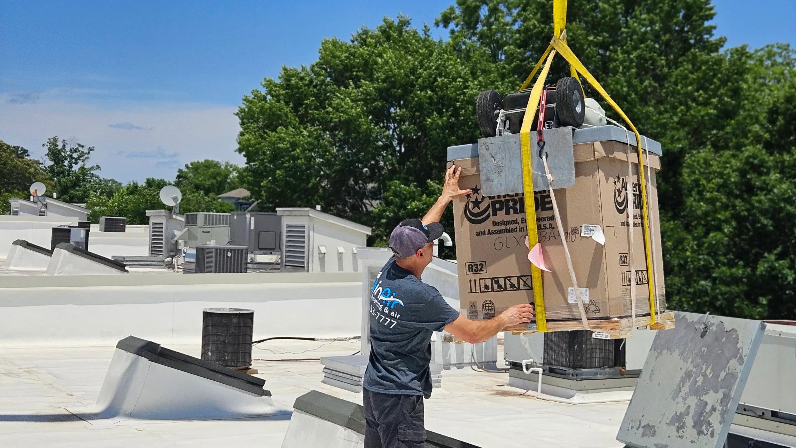 A worker guides a large boxed unit being hoisted by a crane about inair onto a building rooftop under a clear sky, with trees and equipment visible in the background.