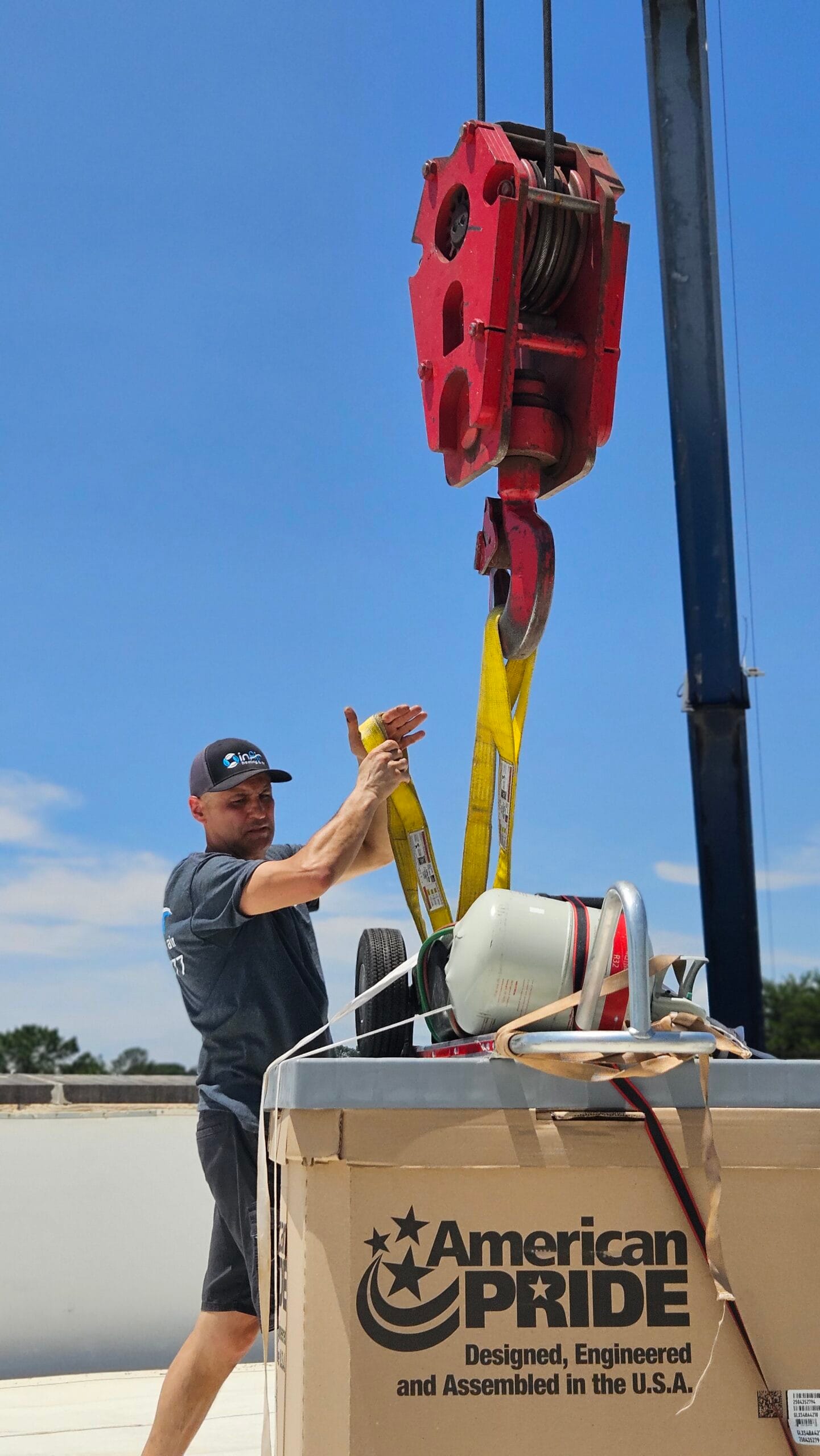A worker guides a large red industrial hook attached to a crane over a box labeled "American Pride" about inair outdoors under a blue sky.