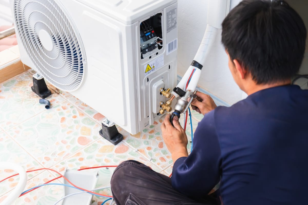 A person installs or repairs an air conditioning unit, connecting refrigerant lines and wiring on a tiled floor, while HVAC repair signs are visible nearby.