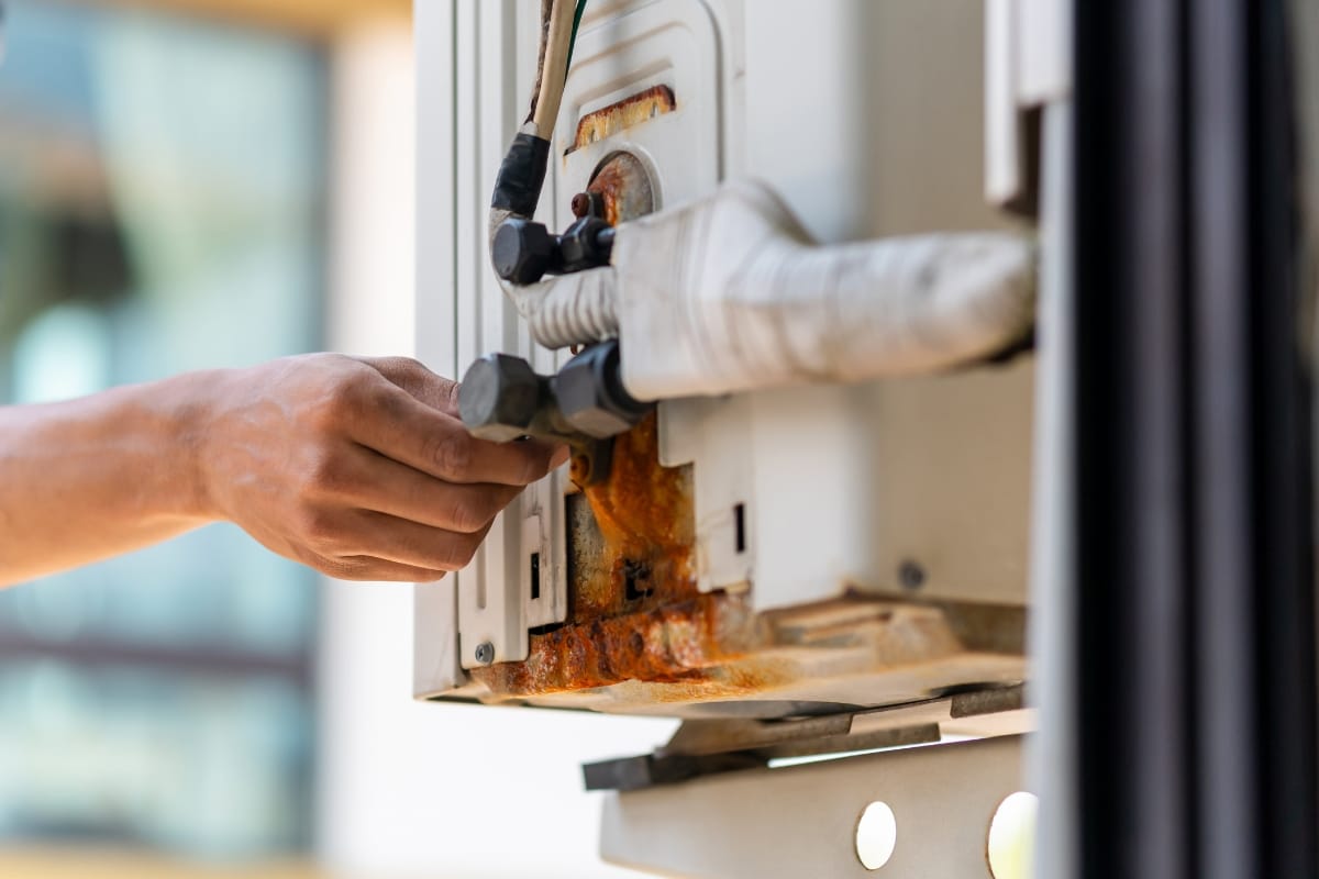 A person’s hand adjusts a fitting on a wall-mounted air conditioning unit, showing visible rust and corrosion around the pipes—common HVAC repair signs.