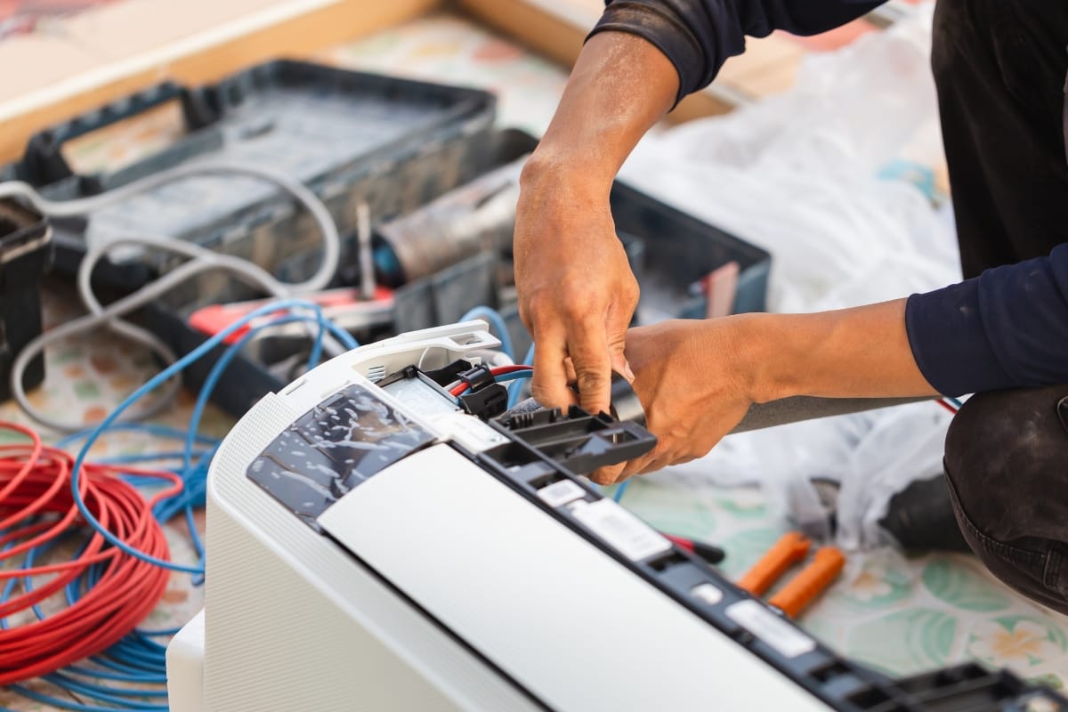Person installing or repairing an air conditioning unit, using tools with electrical wires and equipment visible in the background, carefully following HVAC repair signs for safety and efficiency.