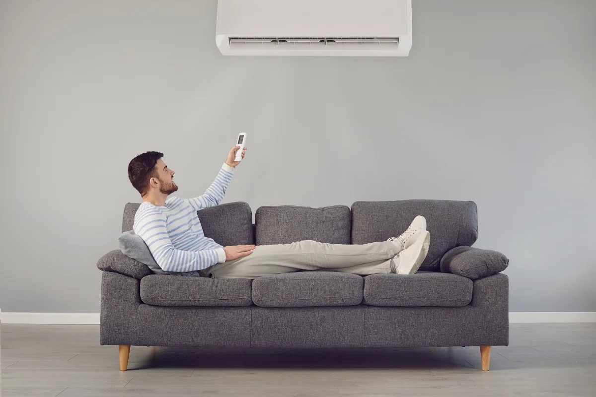 A man lying on a gray sofa points a remote control at the HVAC system mounted on the wall above him.
