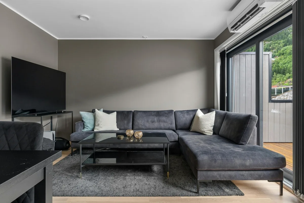 Modern living room with a dark gray sectional sofa, glass coffee table, TV on a stand, and large window overlooking a balcony and greenery outside, complemented by an efficient HVAC system for year-round comfort.