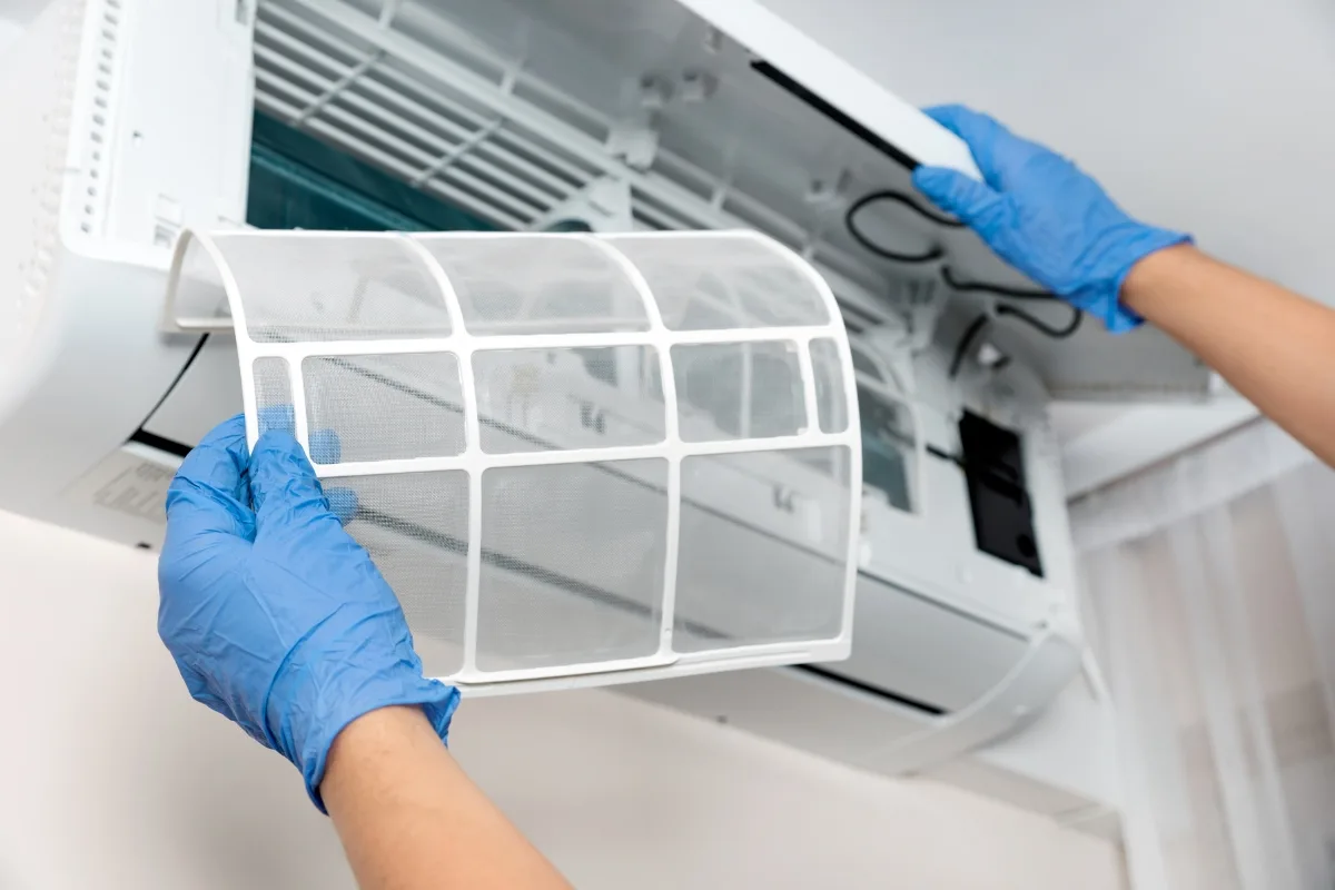 A person wearing blue gloves is removing a mesh filter from an open wall-mounted HVAC system for cleaning or maintenance.