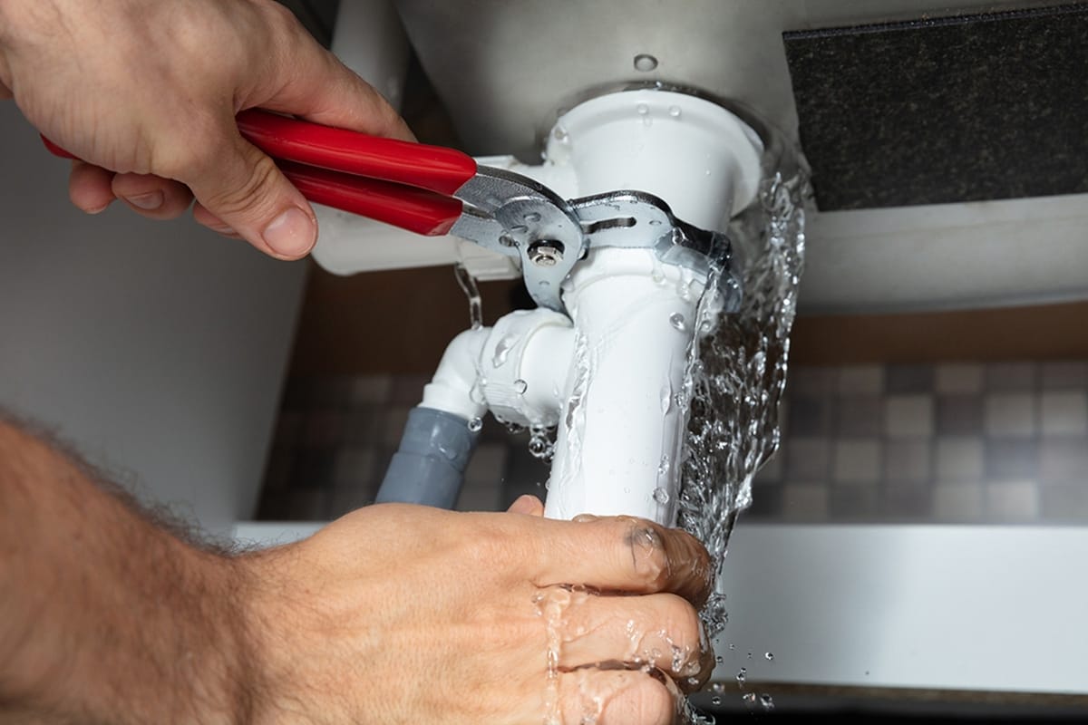 A person uses pliers to fix a leaking white pipe under a sink, water spraying out—just one of the common plumbing problems Georgia homeowners often face.