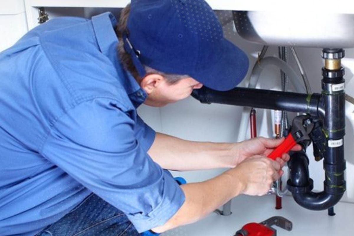 A person wearing a blue shirt and cap uses a pipe wrench to fix plumbing problems under a sink in Georgia.