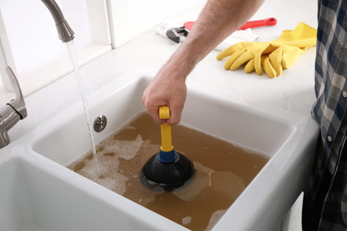 A person tackles plumbing problems in Georgia, using a plunger on a clogged sink filled with dirty water; rubber gloves and a wrench rest on the counter nearby.