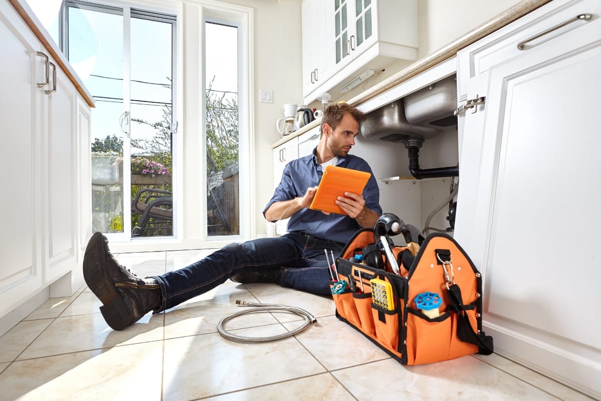 A plumber sits on a kitchen floor inspecting a tablet, with tools and an orange tool bag nearby, while working under a sink to tackle plumbing problems in Georgia.