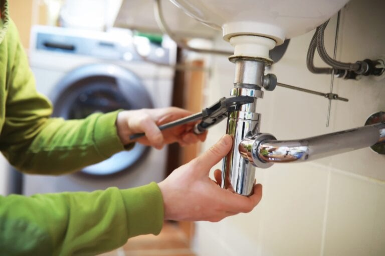 A person uses a wrench to fix plumbing problems under a sink, with a washing machine in the background—typical home maintenance in Georgia.