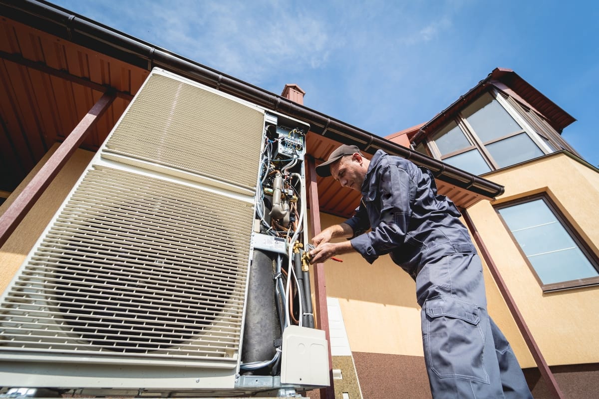 A technician in coveralls performs seasonal HVAC maintenance, repairing an outdoor air conditioning unit attached to a residential building under a clear sky.