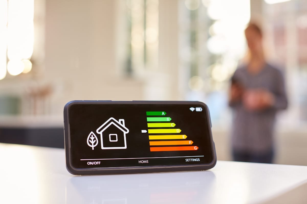A smartphone screen displays a home energy efficiency rating chart, highlighting the importance of seasonal HVAC maintenance, while a person stands in the blurred background holding another device.