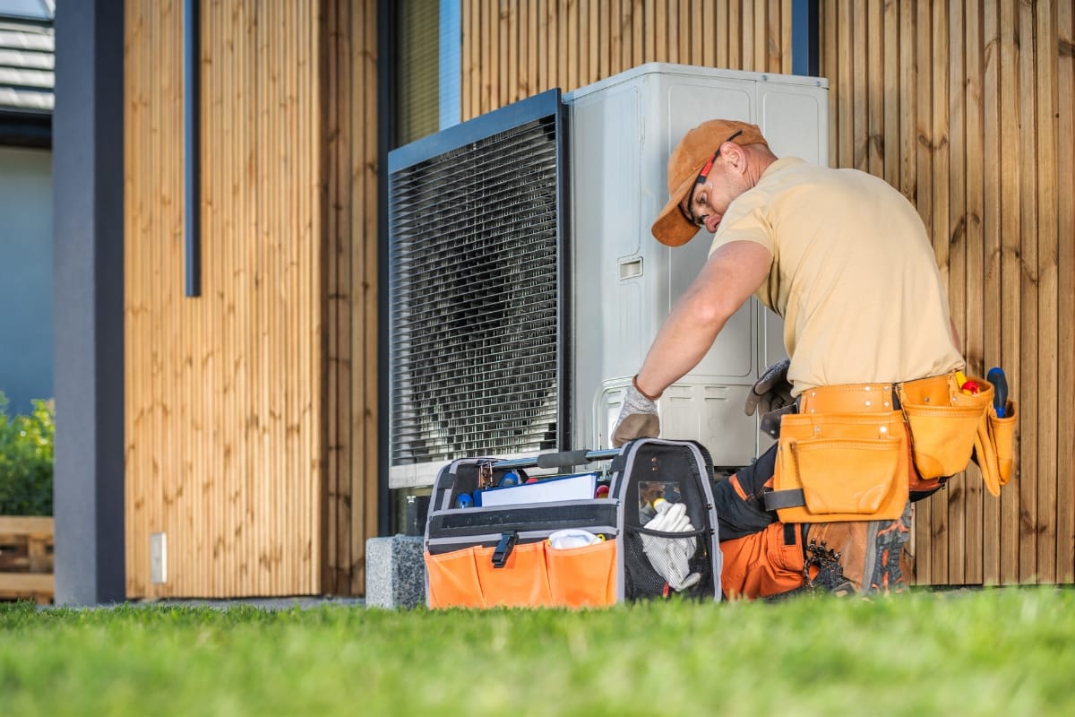 A technician kneeling on grass performs seasonal HVAC maintenance on an outdoor air conditioning unit, with a tool bag open beside him.
