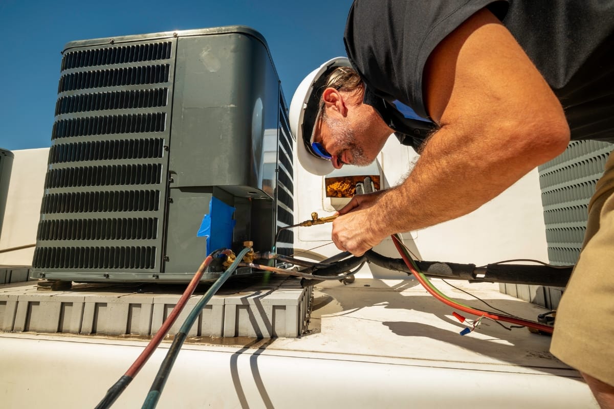 A technician wearing a helmet and sunglasses performs seasonal HVAC maintenance, repairing an outdoor air conditioning unit on a rooftop under a clear sky.
