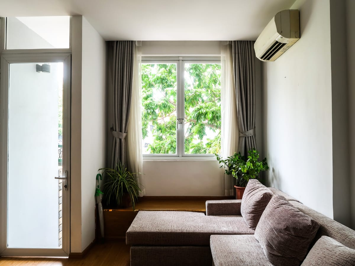 A bright living room with a gray sofa, two potted plants, a large window with curtains, and a ductless mini-split air conditioner mounted on the wall.
