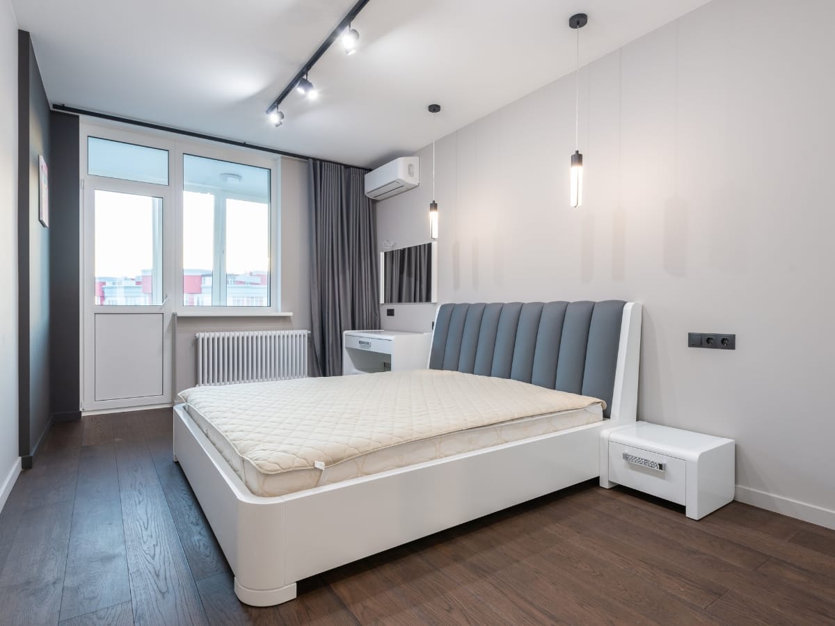 Modern bedroom with hardwood floors, a large bed with a gray and white headboard, pendant lights, a desk, window, neutral-colored walls, and efficient climate control thanks to a ductless mini-split system.
