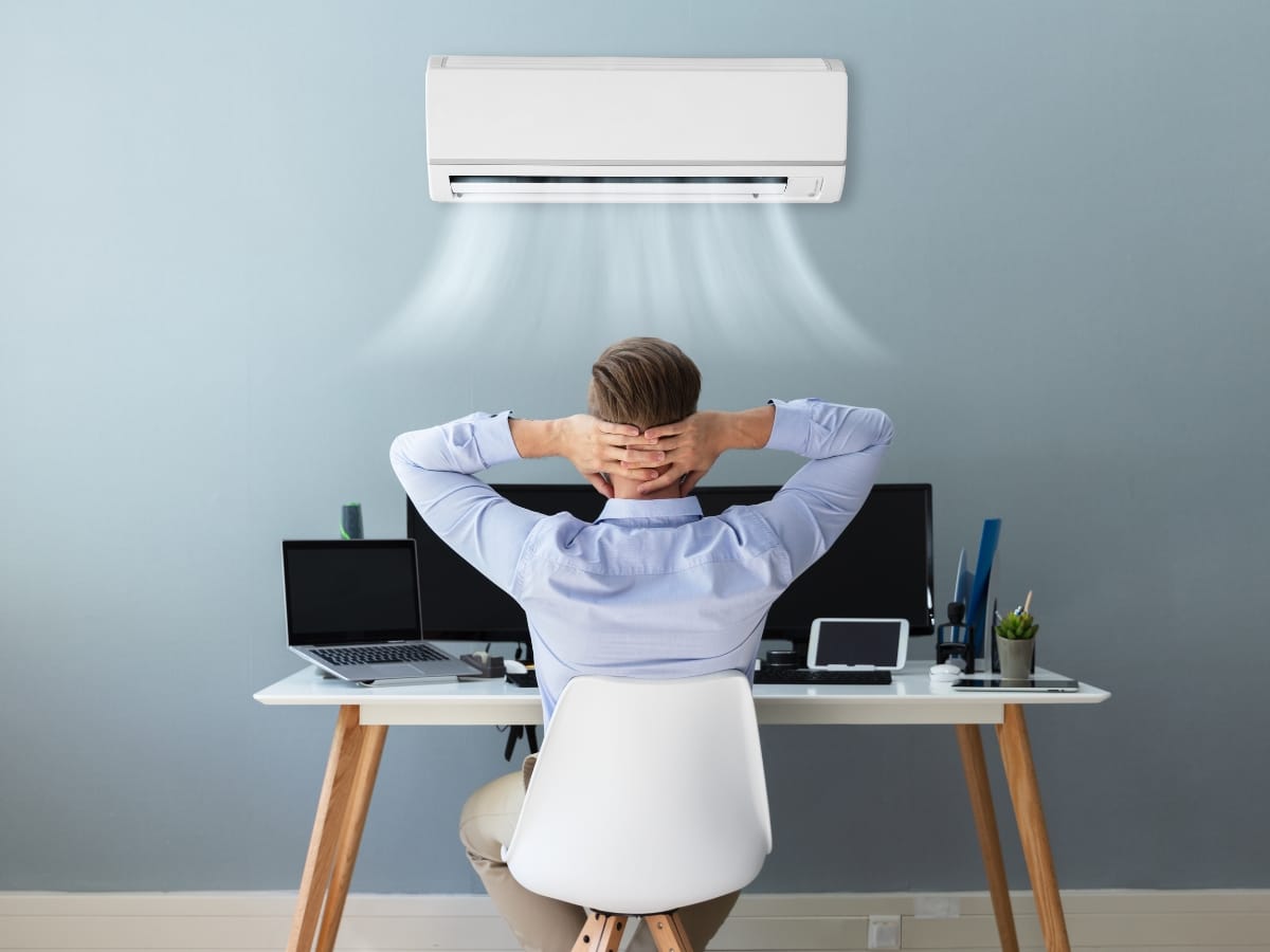 A person sits at a desk with multiple devices, relaxing while a ductless mini-split air conditioner mounted on the wall above blows cool air.