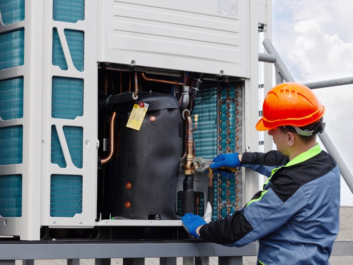 Technician in safety gear performs an HVAC tune-up while repairing or inspecting the components of an outdoor air conditioning or refrigeration unit.