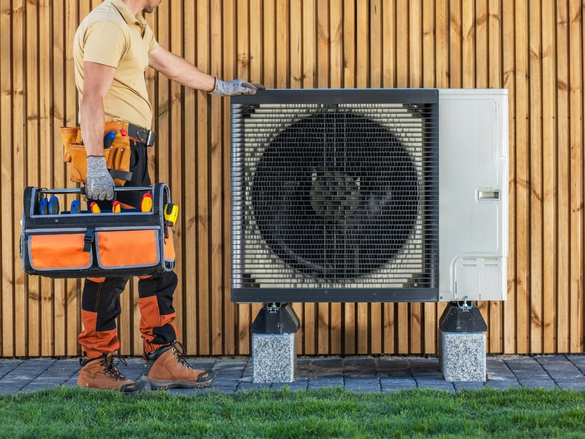 A technician with a tool bag stands next to an outdoor heat pump unit mounted on concrete blocks beside a wooden fence, preparing for an HVAC tune-up.