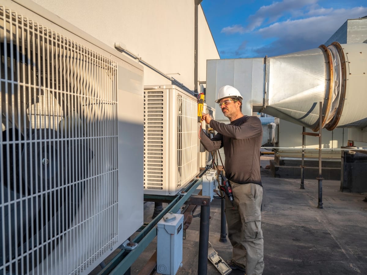 A technician in a hard hat uses a tool to perform an HVAC tune-up or inspect an outdoor unit on a building rooftop.