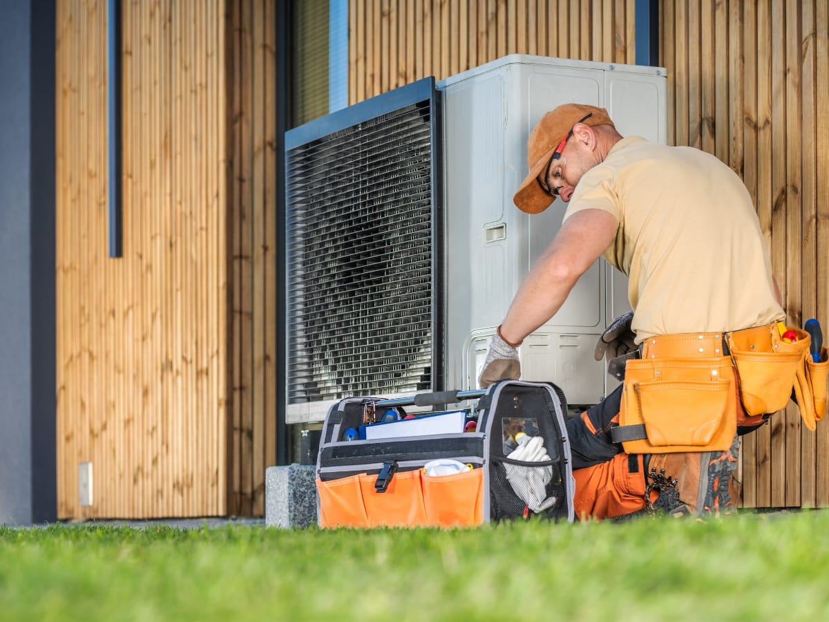 Technician wearing an orange cap performs an HVAC tune-up on an outdoor unit next to a building, with a tool bag placed on the grass nearby.