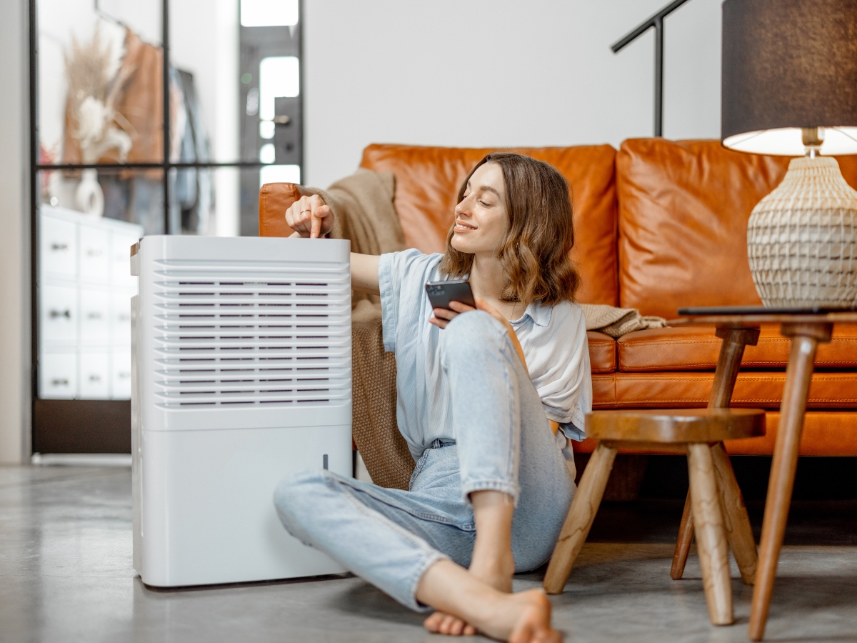 Woman sitting on floor next to brown sofa, adjusting a white air purifier to improve indoor air quality Atlanta residents trust, while holding a smartphone in her other hand.