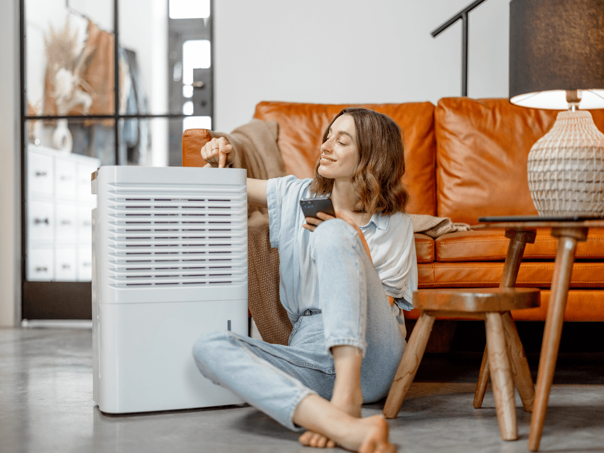 Woman sitting on the floor by a brown leather sofa adjusts a dehumidifier with one hand while holding a remote control, in a modern living room&mdash;demonstrating her commitment to maintaining excellent indoor air quality in Atlanta.